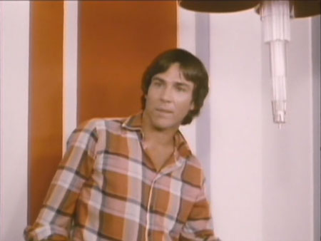 Man in orange plaid shirt stands against orange and white background. His face and figure are brightly lit, with almost no shadowing.