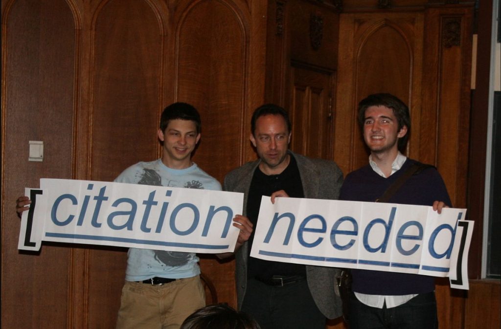 Jimmy Wales and two others hold aloft a Citation Needed sign