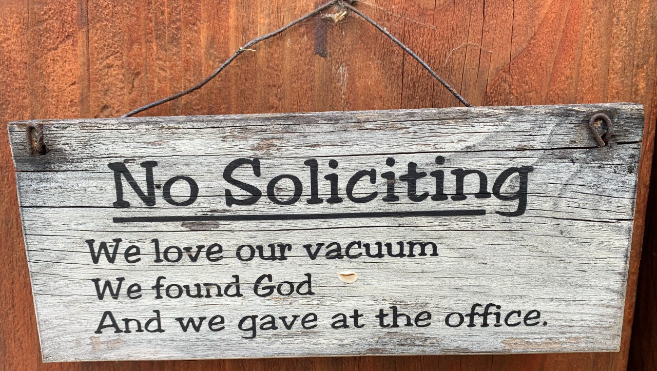 Image of a humorous 'No Soliciting' sign on a door stating, 'We love our vacuum. We found God. And we gave at the office.' The sign playfully dismisses common reasons salespeople, religious groups, and charity organizations visit homes, indicating the householders' desire to avoid unsolicited visits and pitches.